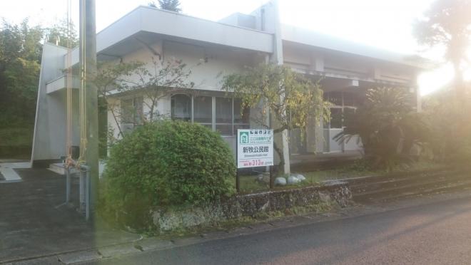 道路側に様々な種類の木が植栽された、白壁で平屋建ての新牧公民館の建物外観の写真