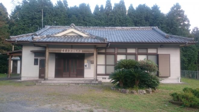 林の手前にある入母屋造りの屋根の平屋建ての手蓑集落公民館の建物外観を植木が植栽された庭から撮影した写真
