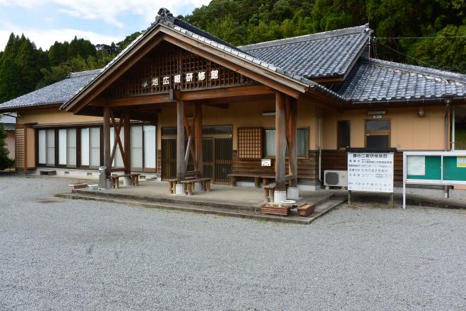 入り口前に三角の屋根の庇がついた、複数のベンチが置かれた広いスペースがある、ベージュ色の壁の厚地公民館の建物外観の写真
