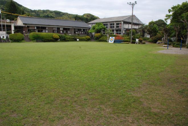 左側に1階建ての校舎と、右側に2階建ての校舎がある旧神殿小学校の建物外観を、ジャングルジムや鉄棒などの遊具が設置されている芝生が広がる校庭側から撮影した写真