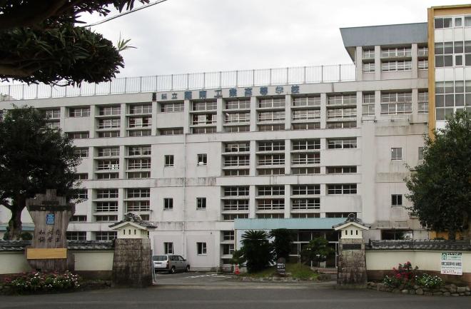 お城を思わせる瓦のある白い塀に囲まれ、門の側にに「鹿児島県立薩南工業高等学校 自主・向学・和協」と書かれたモニュメントがあり、白い外壁の4階建ての校舎の鹿児島県立薩南工業高等学校の外観写真