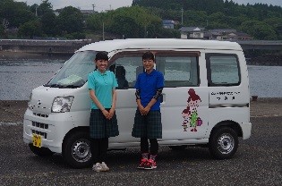 白の「南九州市地域子育て支援センターつみき」の社用車の前に、青と水色のポロシャツを着用したセンタースタッフが2名並んで笑顔で写っている写真