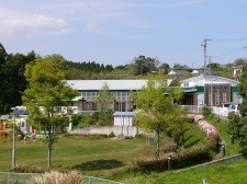 低木の植栽などに囲まれた芝生の園庭から一段高いところに建つ、白っぽい屋根の建物の光栄保育園の写真