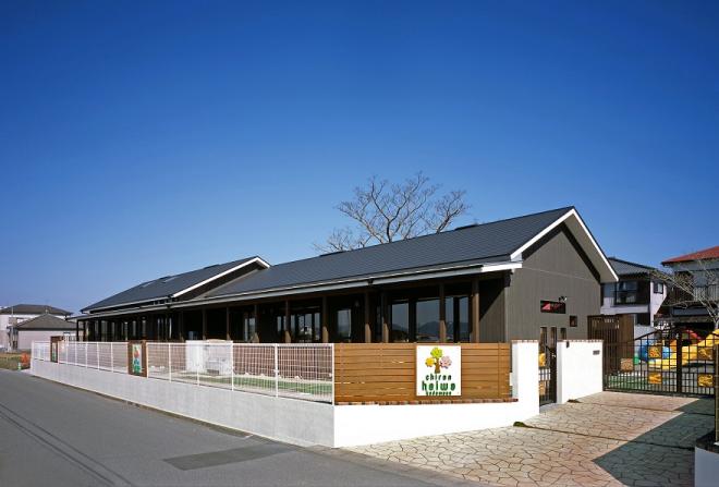 白色のフェンスに囲まれた敷地内に遊具が設置され、グレー色の壁の平屋建てで横長のちらん平和こども園の建物外観の写真