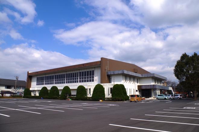 手前には広々とした駐車場が隣接しており、白を基調とし、屋根の上部や壁が一部茶色の外壁をした南九州市知覧体育館の外観写真