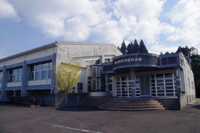 階段を5段上ったところにある玄関の上部に半円形のひさしが施されている、コンクリート調の南九州市知覧武道館の建物外観の写真