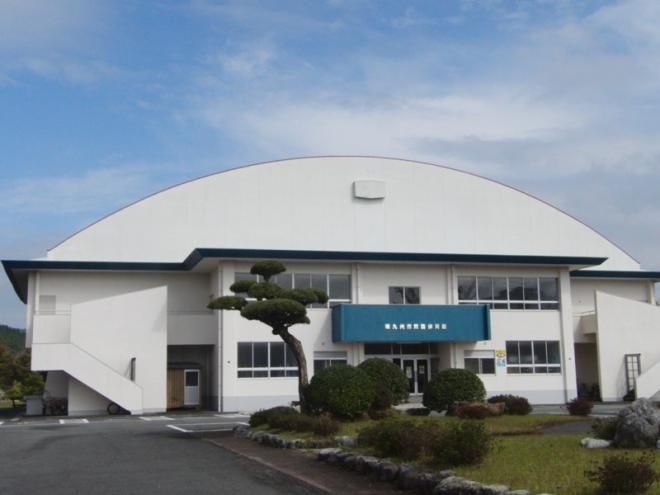 白い外壁の体育館で、建物前には庭木が植えられた芝生の庭が作られ、玄関上に「南九州市諏訪運動公園体育館」と書かれた諏訪運動公園体育館の外観写真