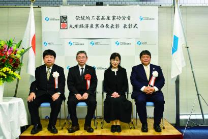 「伝統的工芸品産業功労者 九州経済産業局長表彰 表彰式」と書かれた横断幕が設置されたバックボードの前で、赤や白の胸章をつけた3人の男性と、黒い衣装の女性が並んで座った記念写真