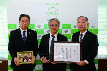 南九州市のバックボードの前で3名のスーツ姿の男性が横に並び、賞状と盾、箱に入った特産品のジャムを手に持ち写した記念写真