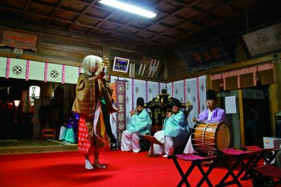 赤い敷物が敷かれた拝殿の中で、笛や太鼓の演奏に合わせ、面をつけて黄色や赤の衣装を身に着けた人物が鈴を持って踊っている写真