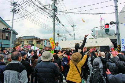 大勢の見物客に囲まれて、1段高いところに茶色の帽子をかぶった男性が立っており、手前では空高く飛んだバナナをつかもうと見物客が手を伸ばしている、バナナのたたき売りの様子の写真