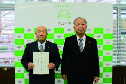 南九州市のバックボードの前で「人権擁護委員」の委嘱を受けた男性2名が並んで立ち、向かって左側の男性が委嘱状を持っている写真