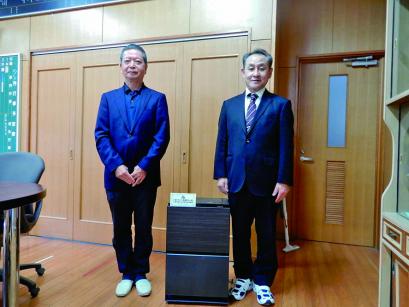 スーツを着て並んで立つ男性2人の間に、太ももあたりまで高さがある、こげ茶色で長方形の空気清浄機が置かれている写真