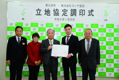 大きく南九州市・株式会社ヨシヤ食品 立地協定調印式 令和5年1月26日と書かれた横長の紙が取り付けられたバックボードの前で、5人の男女が並び、中央の男性2名が一緒に協定書を持っている記念写真