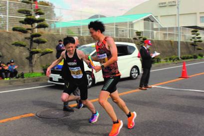 黒いユニフォームの選手から赤に黒の模様が入ったユニフォームの選手にタスキが渡されている場面の写真