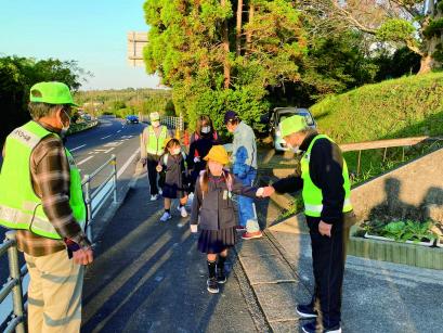 蛍光緑の帽子とベストを身に着けたパトロール隊の男性3人が通学路に立ち、歩いてきた児童や保護者に声をかけている様子の写真