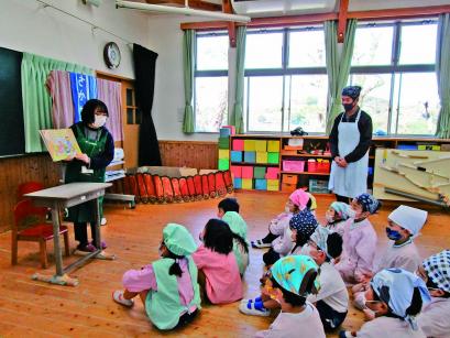 教室の黒板前でエプロンを着た女性が絵本を開き、給食着を着て床に座った子どもたちに見せている写真
