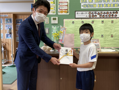 掲示物の貼られた壁の前で、スーツの男性と体操着を着た男子生徒が目録の封筒一緒に持っている写真