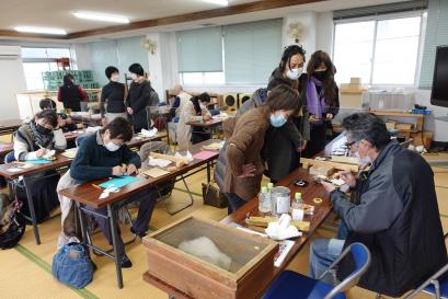 畳敷きの部屋に長机と椅子が複数並べられ、前の席に座った男性から作業の説明を受けている女性たちや机に向かって作業体験をしている女性たちがいる様子の写真