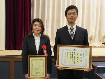 舞台の下に並んで立ち、それぞれ額に入った認定証をカメラに向けて持っている、月野さんと東さんの写真