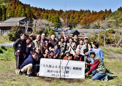 数件立ち並んだ民家の後ろに森が広がっている場所で、植樹祭の参加者たちが3列になって並び、前列の両端の男性が、白地に「「大久保ふるさとの桜」植樹祭 桜がつなぐ絆」と書かれた横断幕を広げて写した記念写真