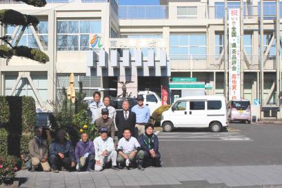 市役所知覧庁舎の門の横に設置された門松の周囲に11人の男性が並び、駐車場の奥にある2階建ての庁舎が入るように写した記念写真