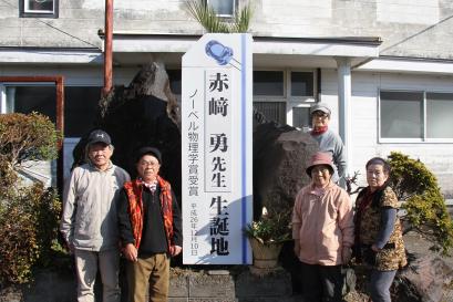 白い壁の建物前の大きな石に設置された、赤崎勇先生生誕地と大きく書かれた立て看板の左右に5名の男女が立っている写真