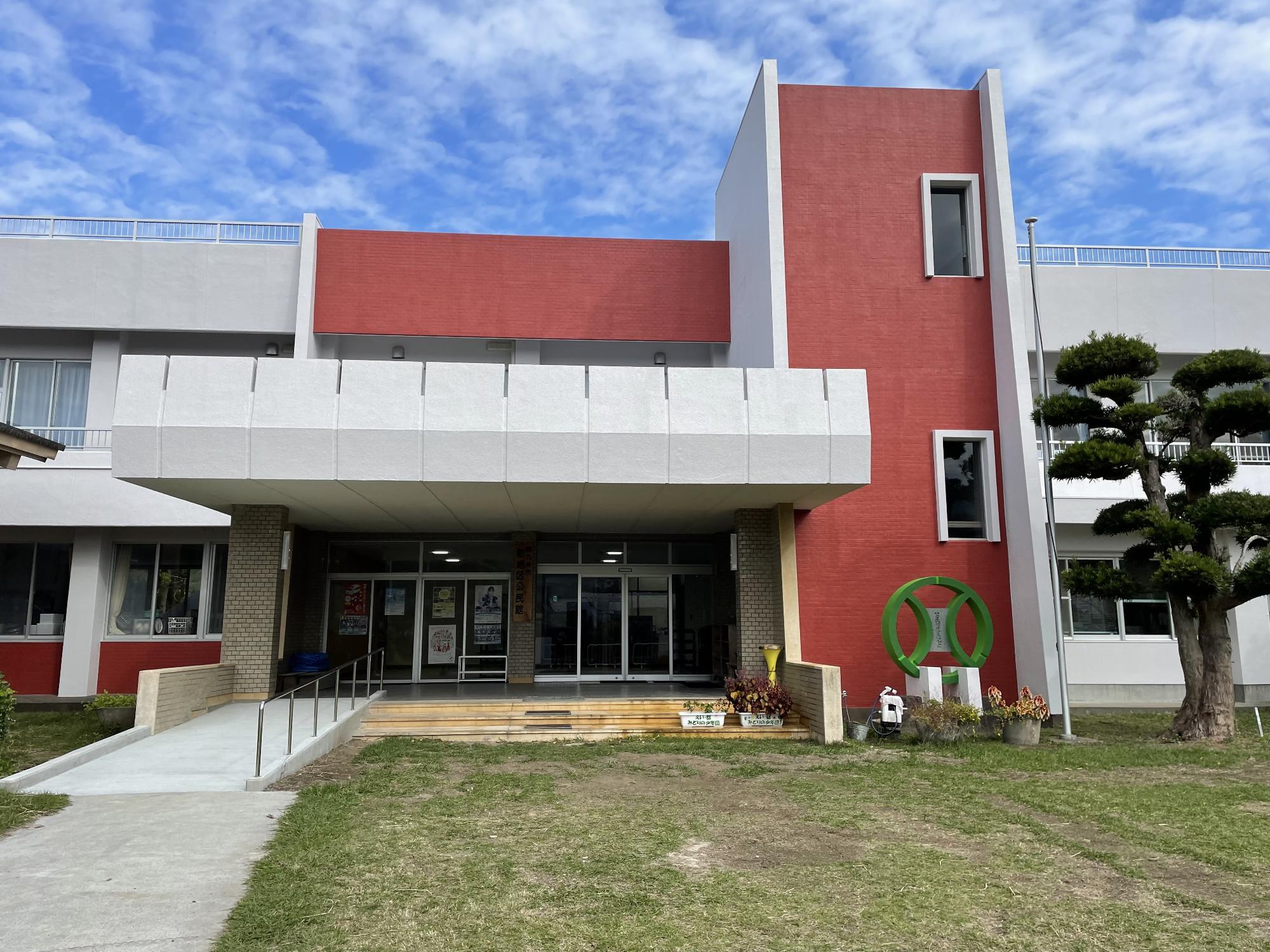 中央部が一部2階建てで縦横にレンガ色のデザインで建物前に芝生の広場が広がっている郡地区公民館の外観写真