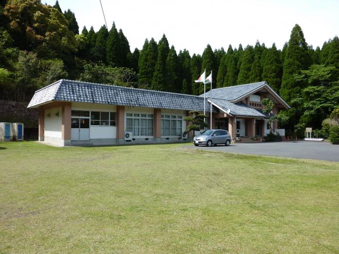 建物の前に旗が2本立っており、緑に囲まれた手蓑地区公民館の外観を芝の広がる場所から撮影した写真