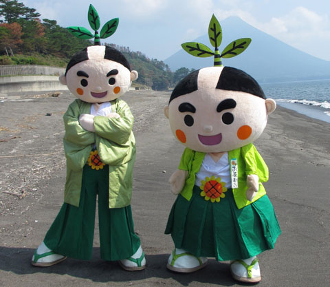 開聞岳が見える浜辺で、腕組をした背の高いお茶むらいと顔の大きい3頭身のお茶むらいが横に並んでいる写真