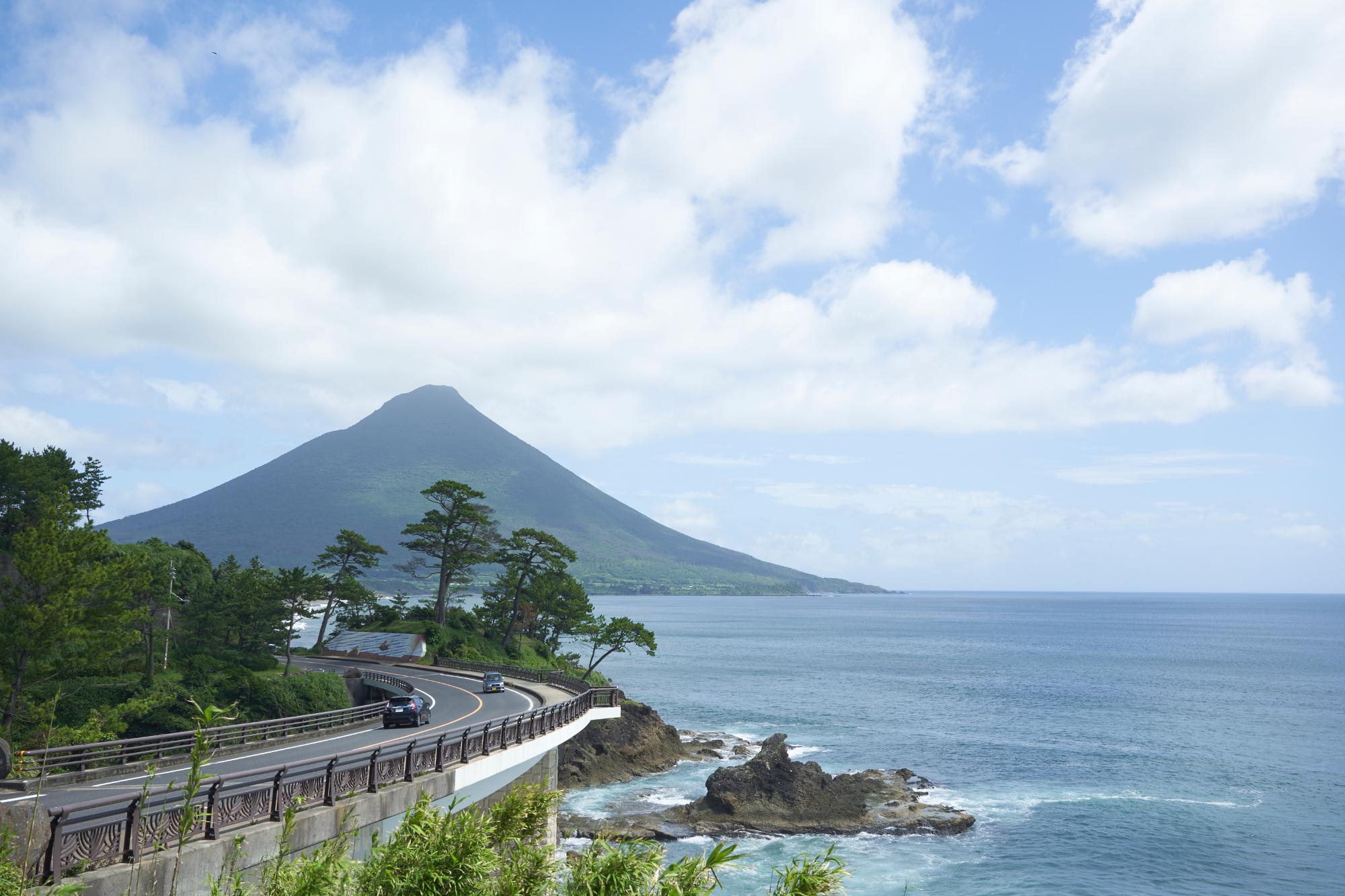 青空の下、右側には青い海が広がり、正面には開聞岳（薩摩富士）、海沿いの道路では車が走っている瀬平公園の写真