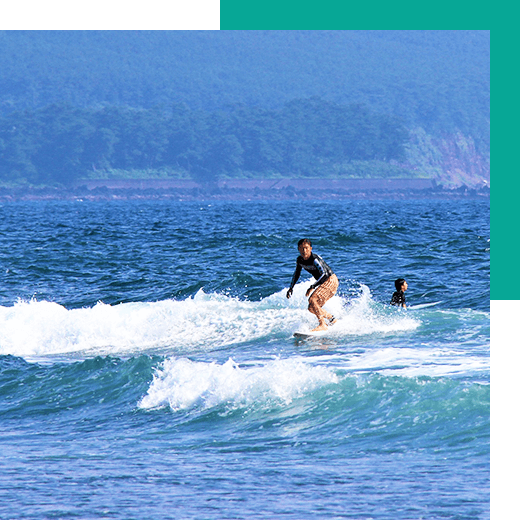 海でサーフィンをしている人が2人。奥には島の沿岸が写っており、自然に恵まれていることが伝わるくらいに木が生い茂っている。